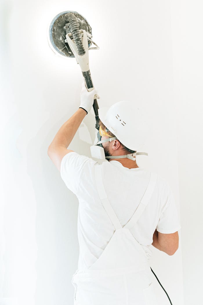 Construction worker smoothing a wall with a power sander, wearing safety gear and white overalls.